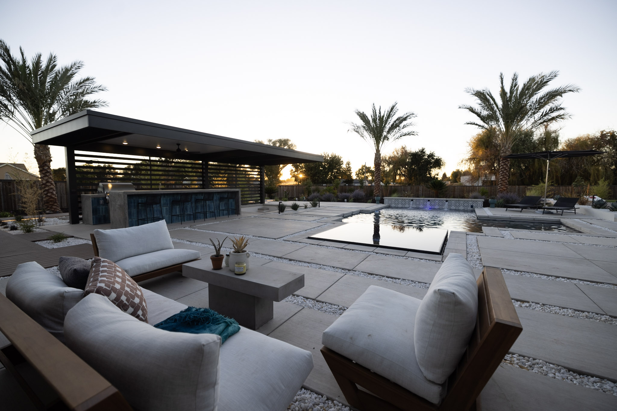 Covered outdoor kitchen and bar beside a rectangular pool at dusk, with large-format paver decking and lounge seating